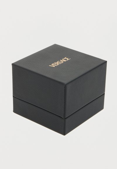 Μαύρο τετράγωνο κουτί με υφή, με το χρυσό λογότυπο VERSACE στο κέντρο στην κορυφή, τοποθετημένο σε απλό ανοιχτό γκρι φόντο.