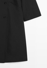 Zwart shirt met korte mouwen, knopen aan de voorkant en een borstzak, afgebeeld van middel tot zoom op een witte achtergrond.