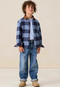 Chemise en flanelle à carreaux bleus et bleu marine avec col en velours côtelé marron, portée sur un t-shirt bleu clair et associée à un jean denim retroussé et des chaussures beiges.