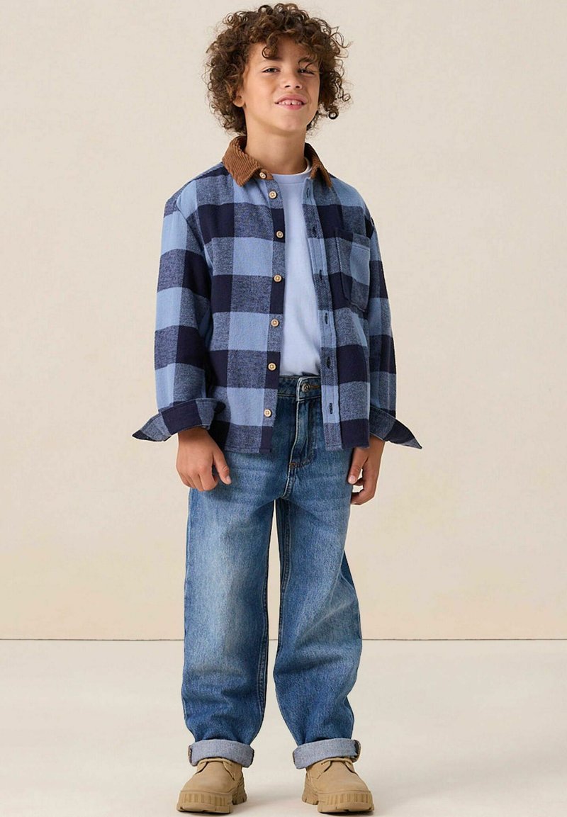 Chemise en flanelle à carreaux bleus et bleu marine avec col en velours côtelé marron, portée sur un t-shirt bleu clair et associée à un jean denim retroussé et des chaussures beiges.