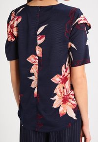 Blusa blu navy con motivo floreale, a maniche corte, caratterizzata da una cucitura sul retro e un tessuto testurizzato. I fiori sono nelle tonalità di rosa e crema.