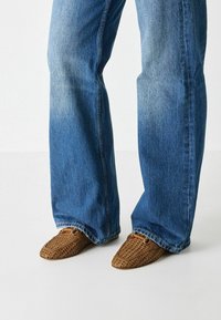 Brown woven loafers with a pointed toe, paired with blue denim flared jeans. The shoes have an open, textured design.