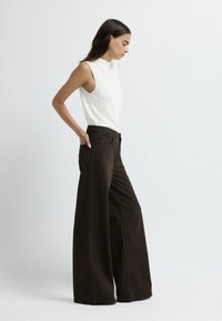 Pantaloni a gamba larga in tessuto marrone scuro con borchie decorative in argento. Abbinati a un top senza maniche bianco aderente, che mette in risalto una silhouette slanciata.