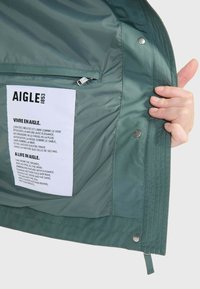 Intérieur d'une veste imperméable verte, avec une étiquette blanche comprenant du texte, une poche zippée et des boutons-pression. Matériau synthétique lisse.