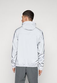 Helle silberne Windjacke mit Kapuze, elastischen Bündchen und Saum. Glatte Textur mit einem schlanken, sportlichen Schnitt. Minimalistisches Design, keine sichtbaren Logos.