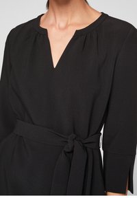 Svart klänning med V-ringning, rynkdetaljer vid axlarna, 3/4-långa ärmar och knytbart midjebälte för justerbar passform.