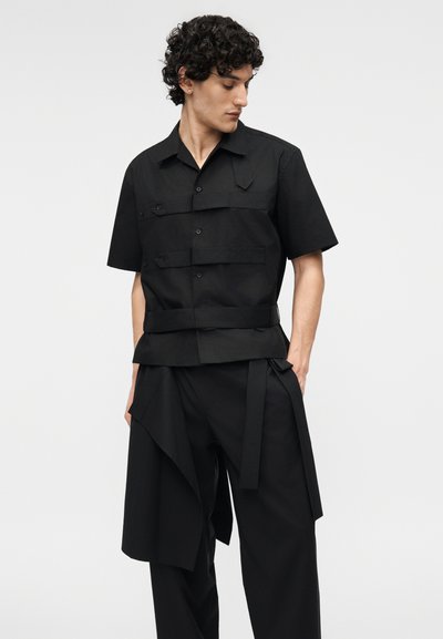 Mladý muž s kudrnatými vlasy, který má na sobě černou krátkosrstou košili se zapínáním na knoflíky a černé kalhoty s detailním vrstveným materiálem, stojí před jednobarevným pozadím.