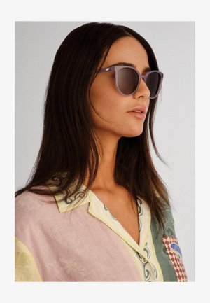 Noor naine, kellel on pikad tumevärvilised juuksed, kannab helelillasid päikeseprille ja pastelseid pleekimisega särk, suunaga paremale tavalise tausta ees.