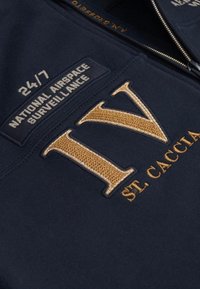 Felpa navy con un grande ricamo dorato "IV" e il testo: "SORVEGLIANZA AEREA NAZIONALE 24/7" su una toppa testurizzata.
