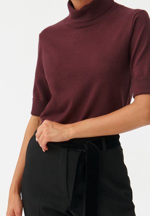 Frau trägt einen bordeauxroten kurzärmeligen Rollkragenpullover, der in eine hochgeschnittene schwarze Hose mit einem schwarzen Samt-Gürtel gesteckt ist.