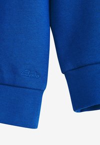 Tessuto in felpa blu con polsini a costine. Presenta un logo Clarks ricamato in modo discreto vicino all'orlo. Materiale morbido e testurizzato.