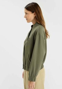 Femme portant une veste zippée olive et un pantalon beige, se tenant de profil contre un fond blanc uni.