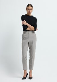 Woman standing with arms crossed wearing black textured top, light gray tailored pants, and black textured high heels on white background.