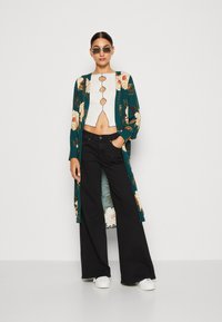 Květinový zelený kimono přes bílé zkrácené tričko s knoflíkovými detaily, spárované s černými širokými džínami a bílými teniskami.