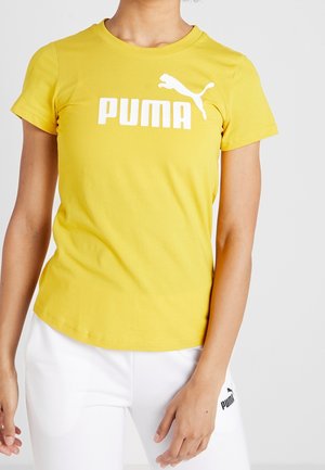 Femme portant un t-shirt jaune Puma avec logo blanc et pantalon blanc avec logo Puma noir sur la cuisse gauche, debout sur un fond uni.