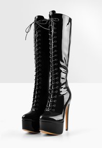 Bottes hautes jusqu'au genou en cuir verni noir, avec un talon aiguille haut, un laçage sur le devant et une semelle plateforme. Texture lisse avec une finition brillante.
