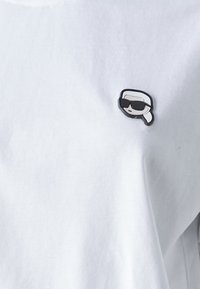 T-shirt bianco in cotone con una piccola spilla in bianco e nero a forma di figura stilizzata con occhiali da sole sulla parte sinistra del petto.