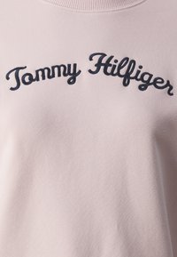 Felpa rosa chiaro con colletto a coste, caratterizzata da un logo ricamato "Tommy Hilfiger" in blu navy scuro sull'abbigliamento. Texture morbida.