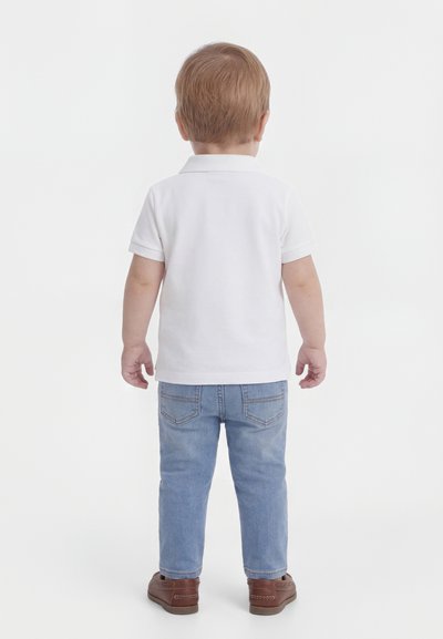 Toddler with light brown hair stands facing away, wearing a white short-sleeve polo, blue jeans, and brown shoes on a white background.
