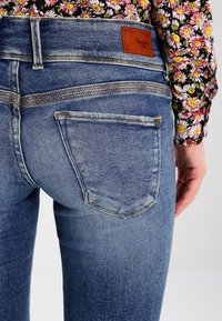 Blå denimjeans med sliten finish, med en brun läderlapp, dubbelsöm och två bakfickor med böjda sömmar.