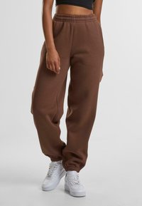 Bruine joggingbroek van zacht materiaal, voorzien van een elastische tailleband en boorden, met zijnaad voor een ontspannen pasvorm. Gecombineerd met witte sneakers.