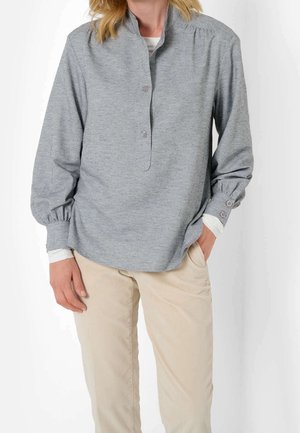 Donna che indossa una camicia a maniche lunghe grigio chiaro con polsini abbottonati e pantaloni beige, in piedi con una mano in tasca contro uno sfondo bianco.