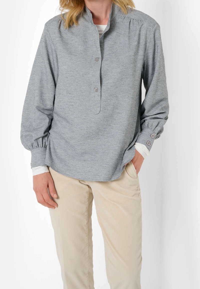 Femme portant une chemise gris clair à manches longues avec poignets boutonnés et un pantalon beige, debout avec une main dans la poche, sur fond blanc.