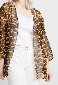 Transparante bruine kimono met luipaardprint, open voorkant en brede mouwen, gedragen over een wit shirt en gecombineerd met een witte broek.