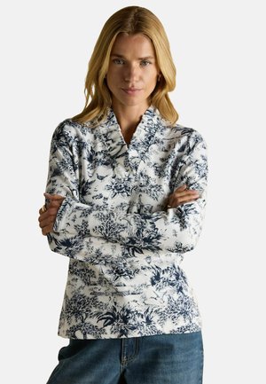 Langarmoberteil mit umgeschlagenem Kragen, verziert mit einem marineblauen Blumenmuster auf weißem Baumwollstoff. Lässiger Schnitt, kombiniert mit blauen Jeans.