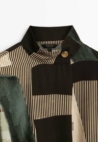 Veste à couches superposées présentant des rayures verticales et horizontales dans des tons de brun et de vert. Tissu texturé avec un col montant et un bouton doré en accent.