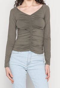 Langärmliges, olivgrünes Oberteil mit V-Ausschnitt und geraffter Textur an der Vorderseite. Kombiniert mit hellblauen Jeans.