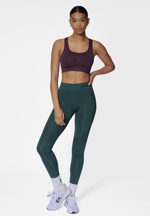 Sportbeha in donkerpaars, gecombineerd met hoge groene leggings met textuurelementen. Model staat in sportschoenen.