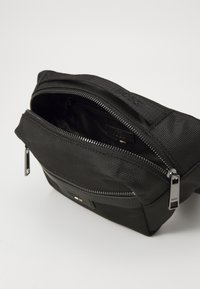 Marsupio nero in nylon con superficie testurizzata, chiusura con zip e accessori in argento. Presenta un'etichetta con logo sul davanti.
