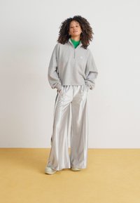 Szary sweatshirt z zamkiem na szyi, z opadającymi ramionami, w zestawieniu z błyszczącymi srebrnymi szerokimi spodniami i jasnym obuwiem, na neutralnym tle.