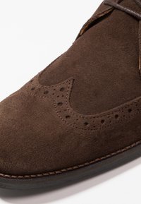 Chaussure habillée en cuir suédé marron avec perforations brogue et surpiqûres détaillées, gros plan sur fond blanc.