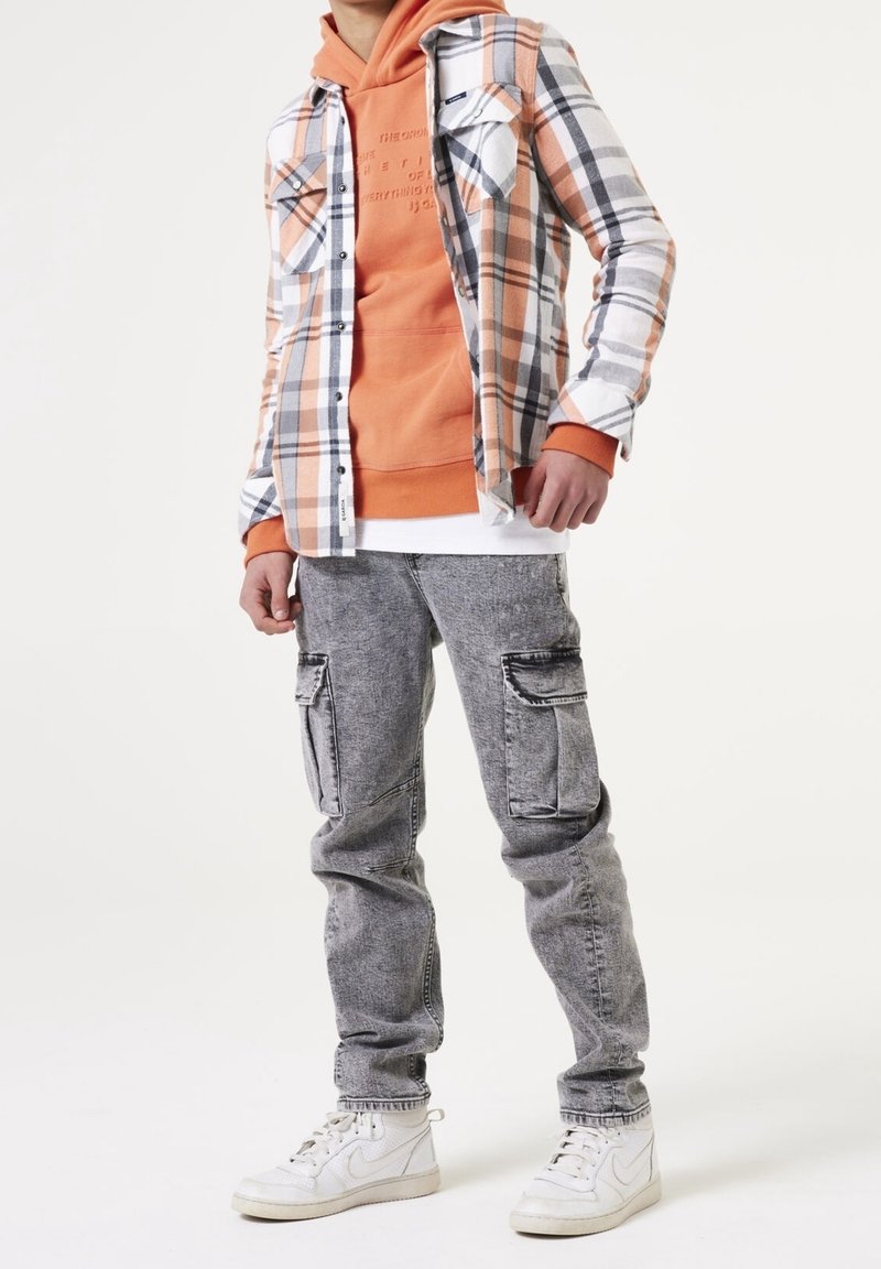 Personne portant un sweat à capuche orange, une chemise à carreaux ouverte, un jean cargo gris et des baskets blanches Nike, debout devant un fond blanc.