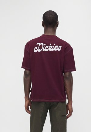 Мъж, носещ бордо тениска Dickies с бял текст на логото отзад и тъмнозелени панталони, показан отзад на едноцветен фон.