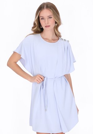 Kobieta o falujących blond włosach, ubrana w jasnoniebieską sukienkę z krótkim rękawem, z guzikami na ramionach i paskiem w talii, pozująca z jedną ręką na biodrze.