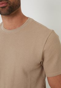 Beige Kurzarm-T-Shirt mit einem strukturierten Waffelstrickmuster und einem runden Ausschnitt. Der Stoff wirkt weich und leicht dehnbar.