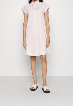Femme vêtue d'une robe-chemise à manches courtes rose clair et de sandales crème avec de larges brides croisées, debout sur un fond uni.