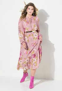 Langes rosa Blumenkleid mit langen Ärmeln, einem volantrücken Saum und einem Schlüsselloch-Ausschnitt. Kombiniert mit rosa Stiefeletten. Ein goldener Gürtel betont die Taille.