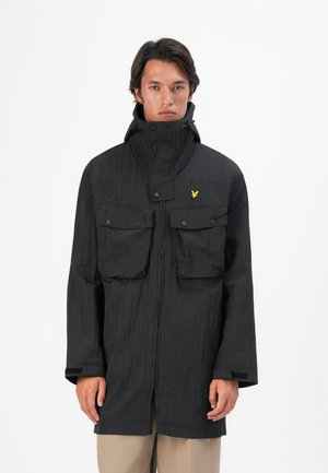 Mann i svart hettejakke med store knappede brystlommer og beige bukser, som står mot en ensfarget hvit bakgrunn.