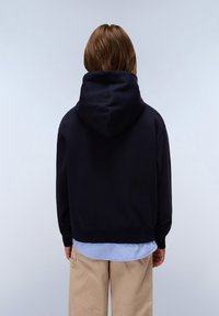 Felpa blu navy con cappuccio regolabile, caratterizzata da un ampio cappuccio e polsini a costine. Indossata sopra una camicia azzurro chiaro e pantaloni beige.