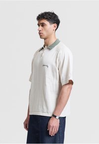 Homme aux cheveux bouclés foncés portant un polo crème à manches courtes avec un col vert et un pantalon bleu foncé, debout devant un fond clair uni.