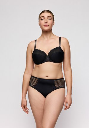 Vrouw staand, gekleed in een bijpassende zwarte kanten BH met dubbele bandjes en high-waist slip met kanten zijpanelen, tegen een effen achtergrond.