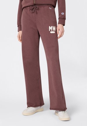 Persona che indossa pantaloni larghi marroni con logo "MW" e scarpe da ginnastica bianche, in piedi contro uno sfondo chiaro e uniforme.