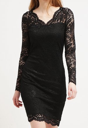 Kobieta ubrana w dopasowaną czarną koronkową sukienkę z długimi rękawami oraz wykończeniem z falbanką na dekolcie i dole, stojąca na jednolitym tle.