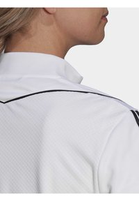 Weißes Poloshirt aus strukturiertem Stoff, mit schwarzen Akzenten an den Schultern und einem hohen Kragen mit leichter Transparenz.