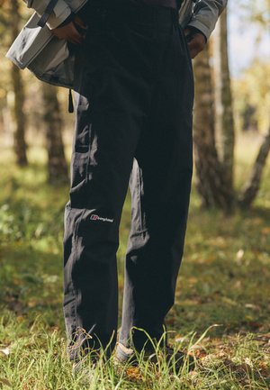 Black outdoor pants by Berghaus, featuring a relaxed fit, durable fabric, a logo on the thigh, and a textured surface, set in a natural environment.