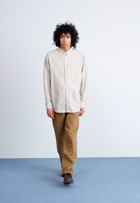 Langærmet button-up skjorte i lys beige med lodrette striber, parret med brune bukser. Bemærkelsesværdig detalje inkluderer en brystlomme.